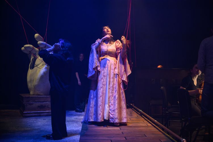 Opera singer in pink silk floor length gown with long draping sleeves holding onto the red string in the set with production team busy working around the singer